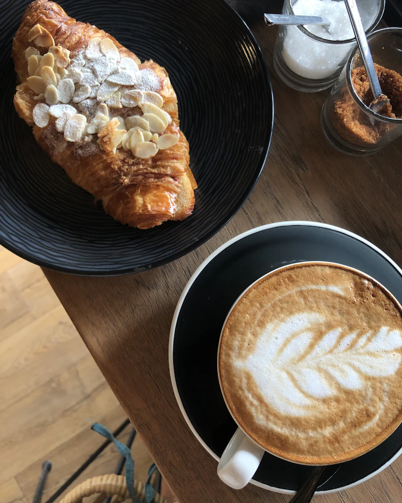 Cappuccino mit Latte Art neben einem Mandelcroissant auf einem Holztisch